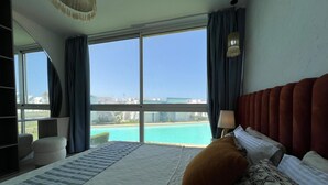 A bedroom with a large window, a bed with patterned pillows, a red headboard, and a side table with a lamp. - Fouka North Coast CL3-CH18-01 (Marsa Matrouh)