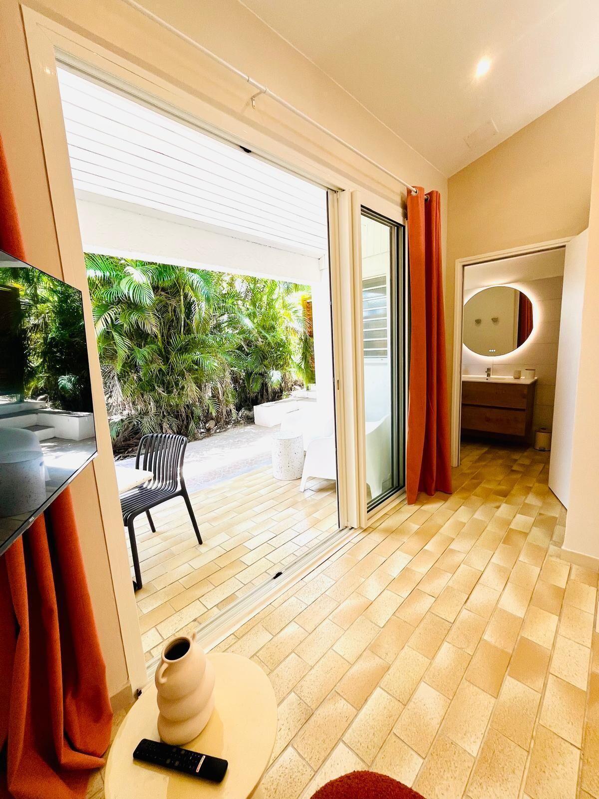 A modern hotel room with a sliding glass door to a patio, a round mirror, a small table with a vase and remote control, and a checkered floor.