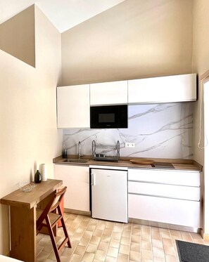 A compact kitchen with white cabinets, a microwave, a small refrigerator, and a wooden table with a chair. - Studio Ti'Bwa - Terrace 2 pers, 3mn from Anse Mitan beach (Les Trois-Îlets)
