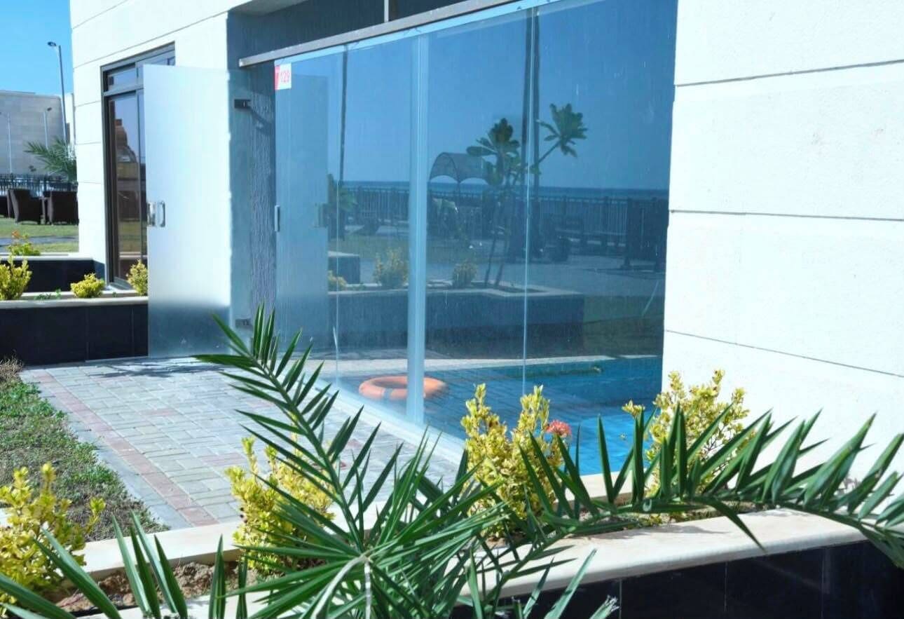 Un edificio moderno con puerta de vidrio, área de piscina con plantas y vista despejada a la playa.