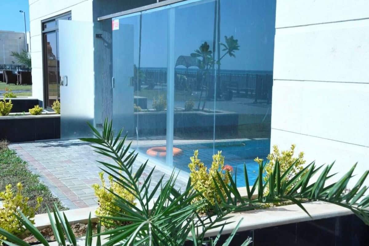 Un edificio moderno con puerta de vidrio, área de piscina con plantas y vista despejada a la playa.