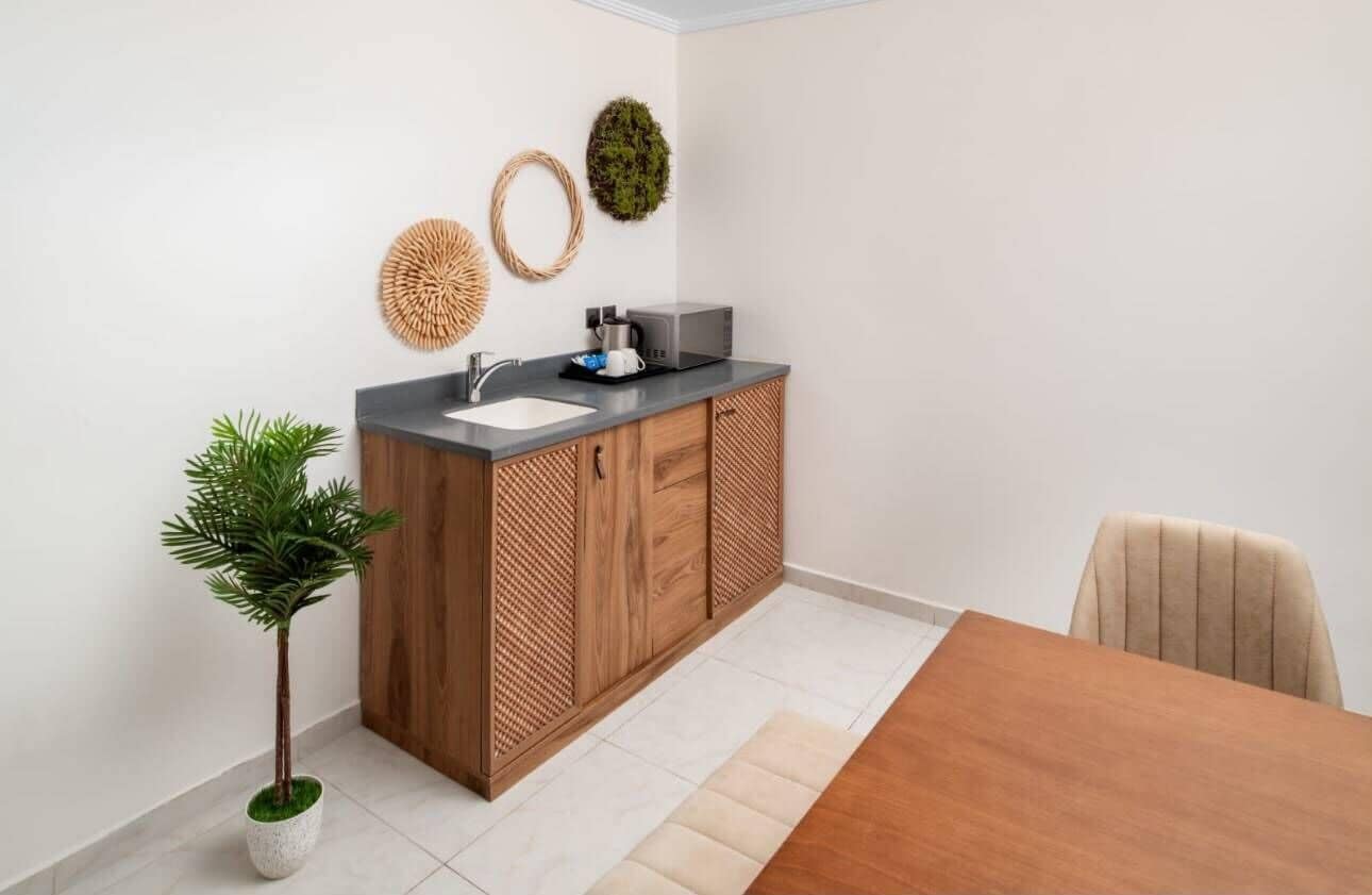 Una cocina compacta con pileta, microondas y muebles bajos de madera. Hay una planta en maceta y una mesa de comedor con sillas.