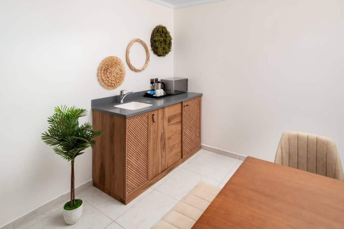 Una cocina compacta con pileta, microondas y muebles bajos de madera. Hay una planta en maceta y una mesa de comedor con sillas.