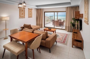 A modern living room with a sofa set, a wooden dining table, and chairs, a small side table, a floor lamp, and a view of the sea through a large sliding glass door.