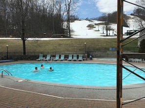 Ein Schwimmbad mit darin badenden Personen, umgeben von einer Ziegelterrasse, und eine Skipiste im Hintergrund.