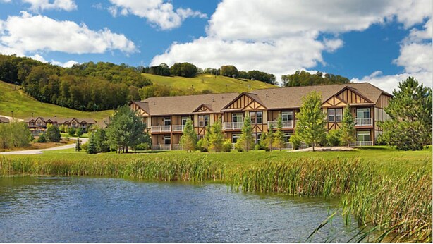 A hotel with multiple buildings, a pond in the foreground, and a hilly landscape in the background. - Enjoy Labor Day weekend in a beautiful 2 BR fully equipped luxury suite. (Boyne Falls)