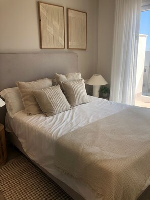 Uma cama com lençóis brancos, cabeceira e várias almofadas. Há uma luminária em uma mesa de cabeceira e uma janela com cortinas.