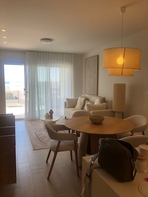 Ein modernes Wohnzimmer mit einem runden Holztisch, weißen Sofas und einem großen Fenster mit durchsichtigen Vorhängen.
