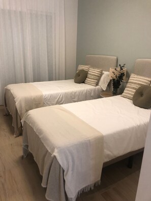 Duas camas de solteiro com roupa de cama branca e cabeceiras bege, uma pequena mesa de cabeceira com um vaso de flores, e uma janela com cortinas leves.