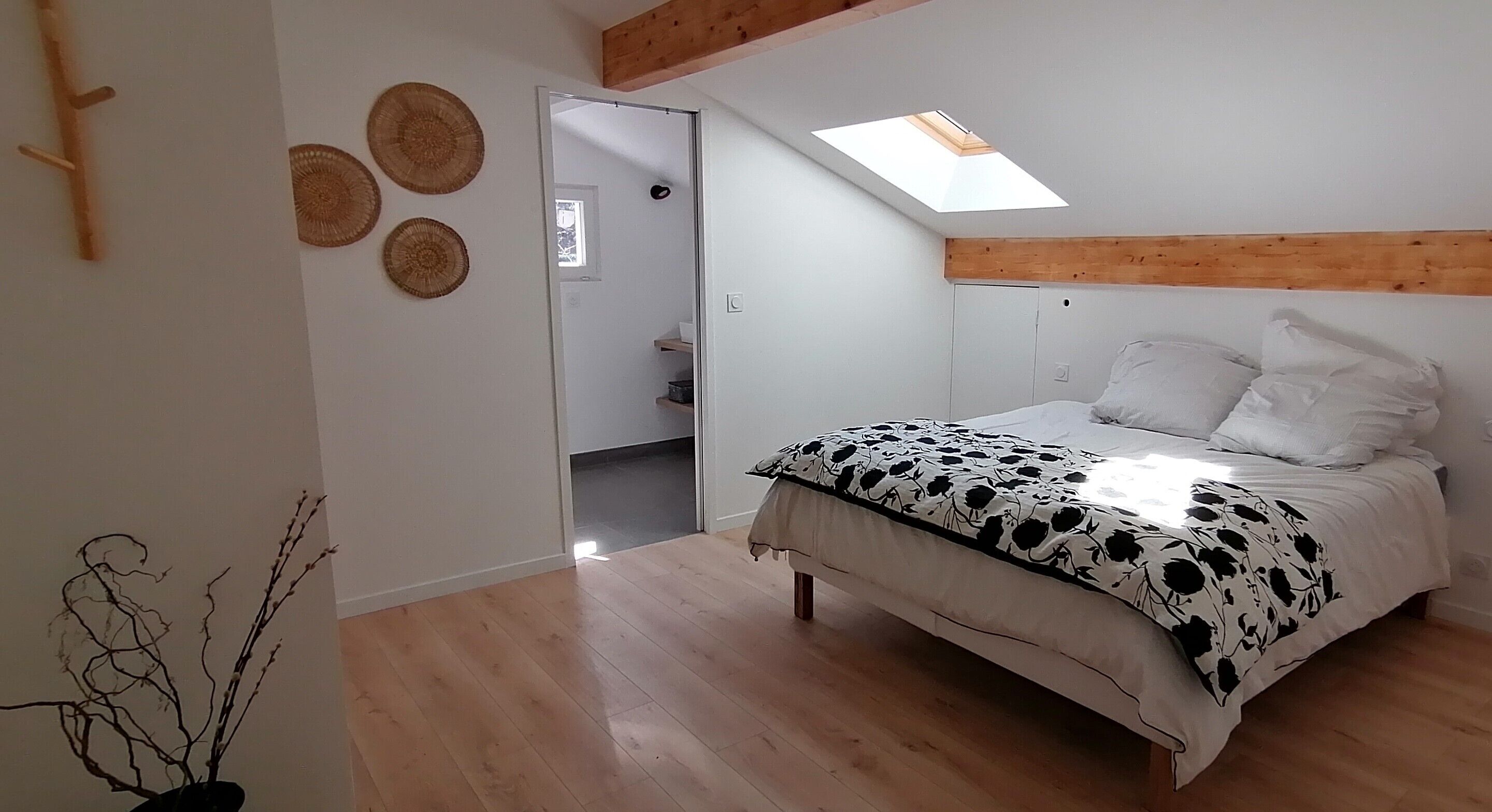 A bedroom with a bed, a skylight, a wooden beam ceiling, and a door leading to a bathroom.