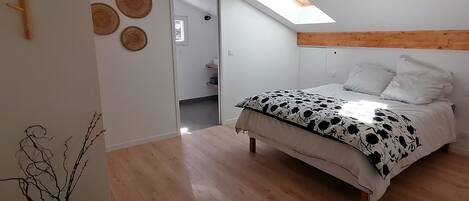A bedroom with a bed, a skylight, a wooden beam ceiling, and a door leading to a bathroom.