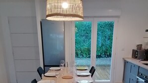 A dining area with a long table set for a meal, a kitchen area with modern appliances, and a view of greenery outside. - Villa Oïhana 4*, 5 suites, Heated pool, Beach 5 km (Labenne)