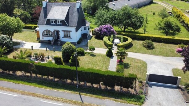 Une grande maison avec un toit à forte pente, entourée de jardins bien entretenus et d’une allée pavée.