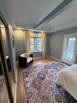 Un dormitorio con cama, una silla, una ventana con cortinas, una puerta con persianas y una alfombra estampada.