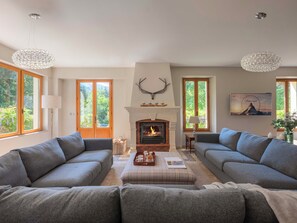 Ein Wohnzimmer mit zwei grauen Sofas, einem Kamin und einem großen Fenster mit Blick auf Grünflächen.