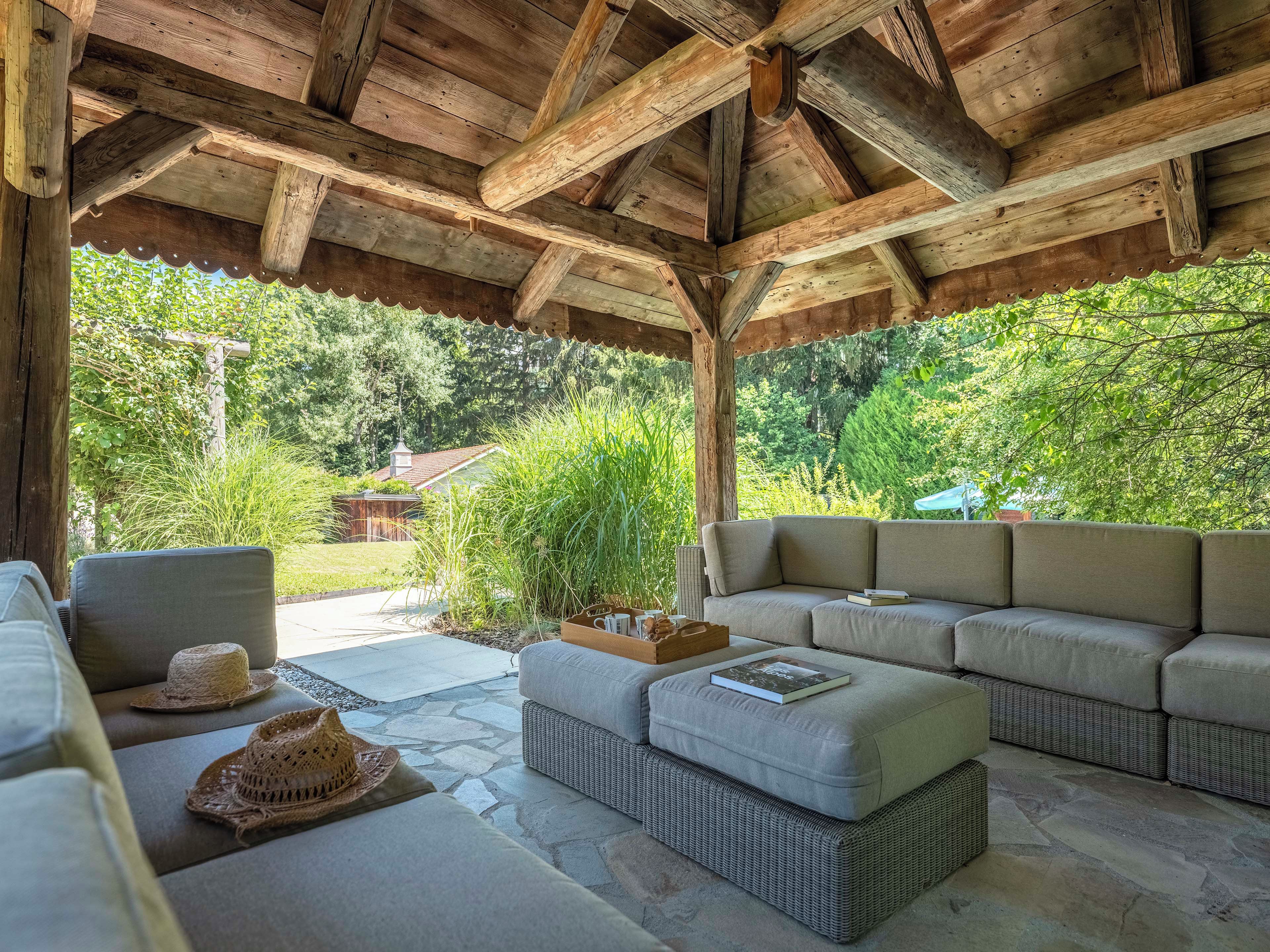 Ein überdachter Außenbereich mit Steinboden, Holzbalkendecke und Blick auf einen Garten sowie ein Haus im Hintergrund.