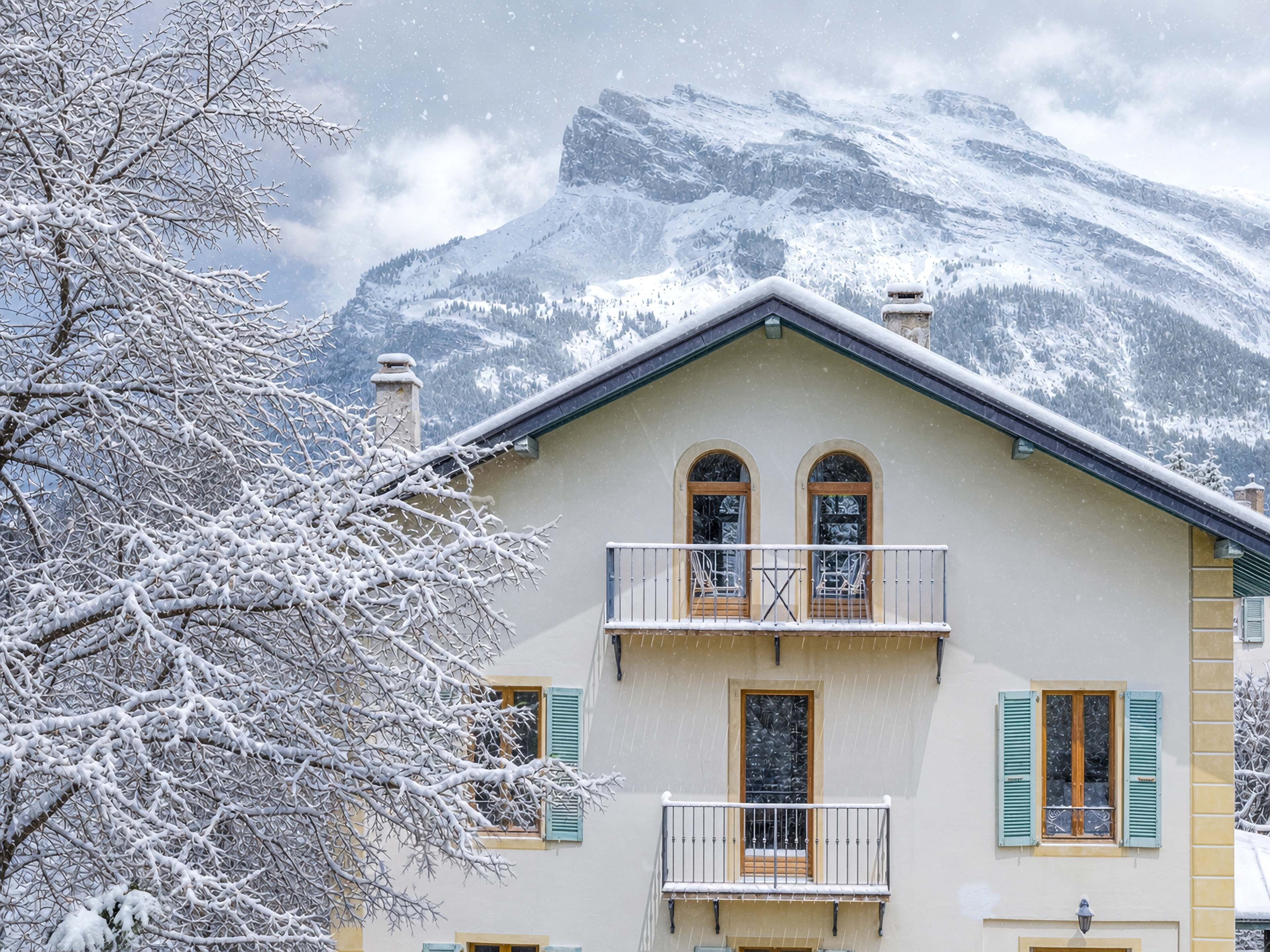 A two-story building with a balcony, a snow-covered mountain in the background, and a tree with snow-laden branches.
