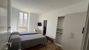 Een moderne slaapkamer met twee bedden, een televisie, een kledingkast en een raam met uitzicht op gebouwen.