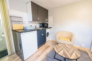 A compact kitchen with a microwave, sink, and washing machine. There's a chair with a cushion and a herringbone-patterned coffee table.