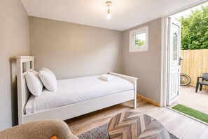 Ein Zimmer mit einem Einzelbett, Holzboden, einem kleinen Fenster und einer Tür zu einem Außenbereich.