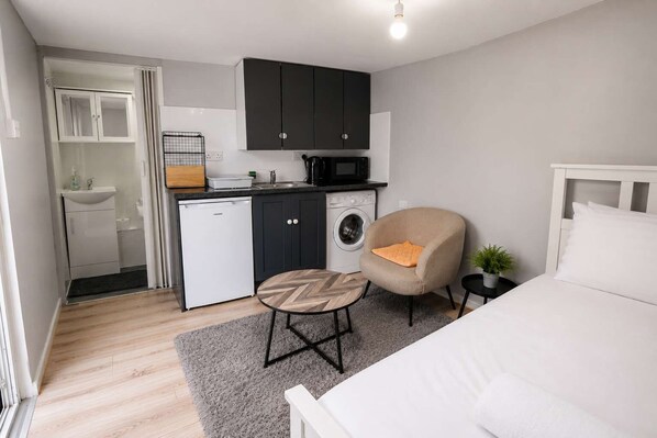 Ein kleines Hotelzimmer mit einem Bett, einem runden Tisch, einem Sessel, einer Waschmaschine und einer Küchenzeile.