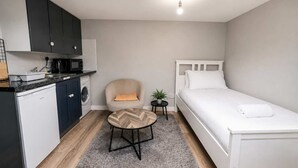 Ein kleines Zimmer mit Küchenzeile, einem Einzelbett, einem Sessel und einem kleinen Tisch.