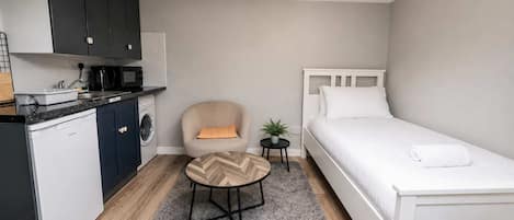 A compact room with a kitchenette, a single bed, a chair, and a small table.