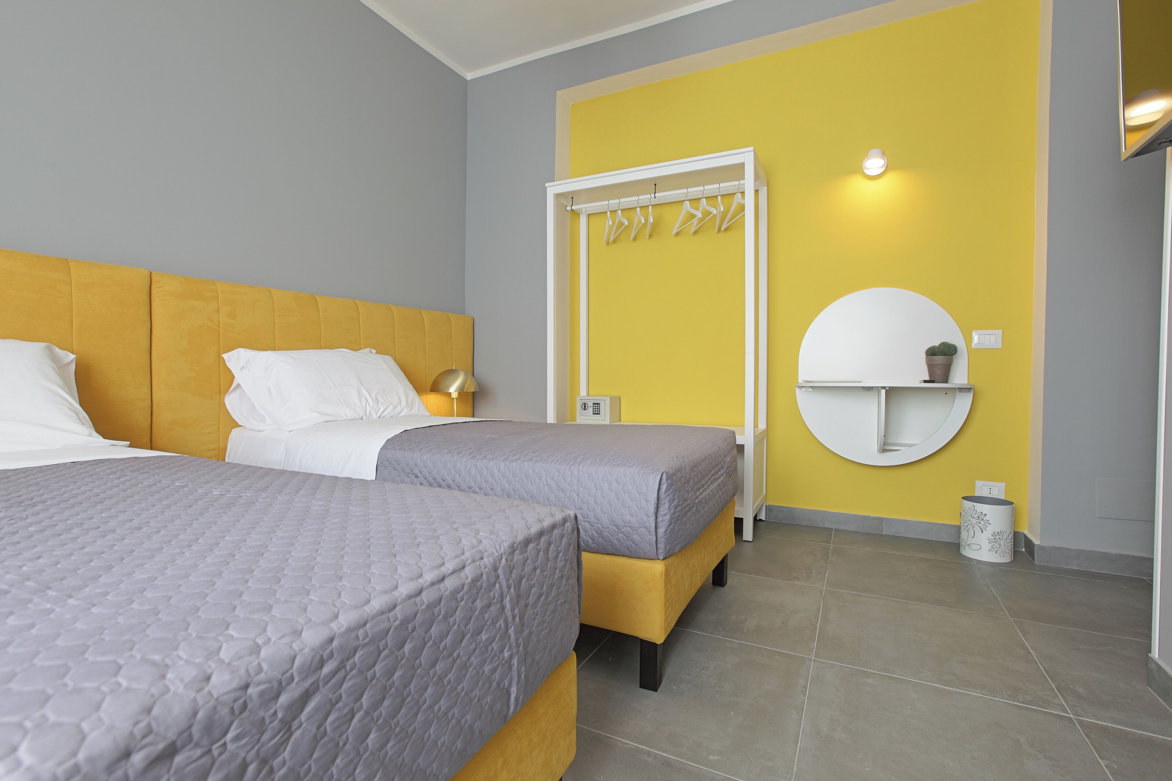 Quarto de hotel com duas camas, parede amarela de destaque, espelho redondo e porta-toalhas fixado na parede.