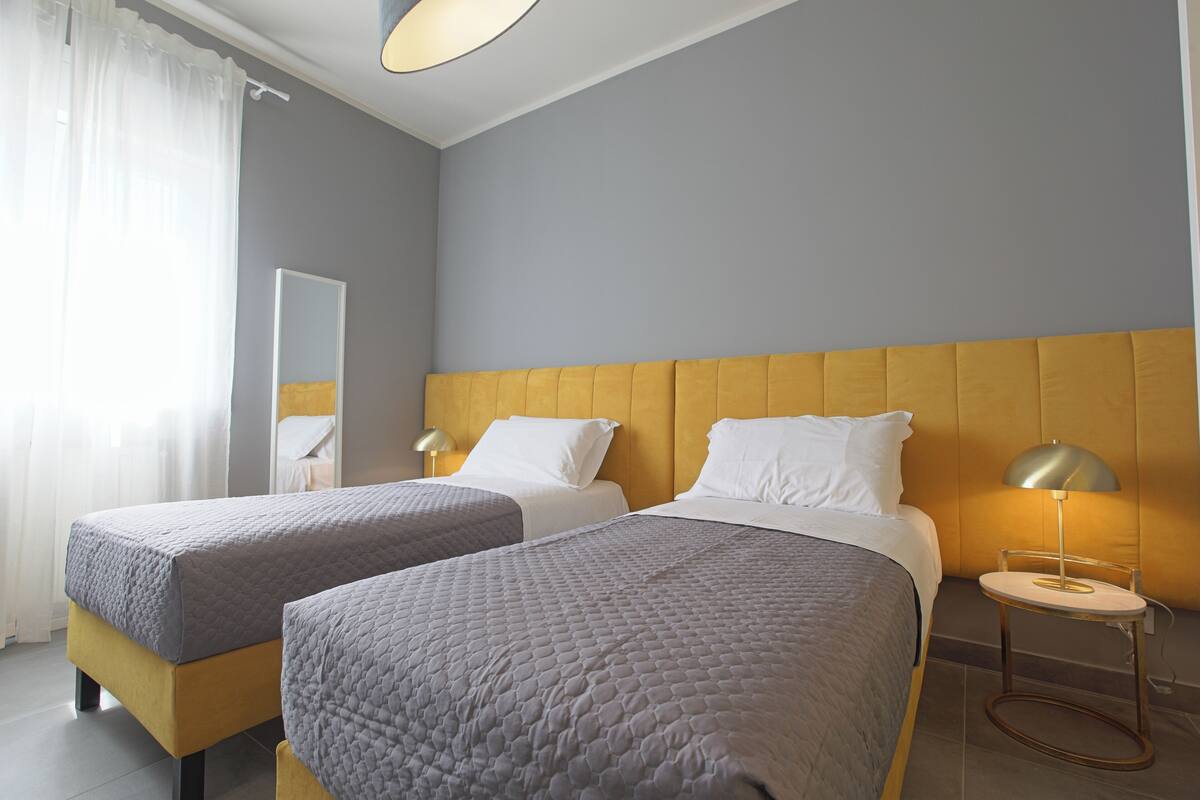 A hotel room with two beds, a bedside table with a lamp, a mirror, and a window with curtains.
