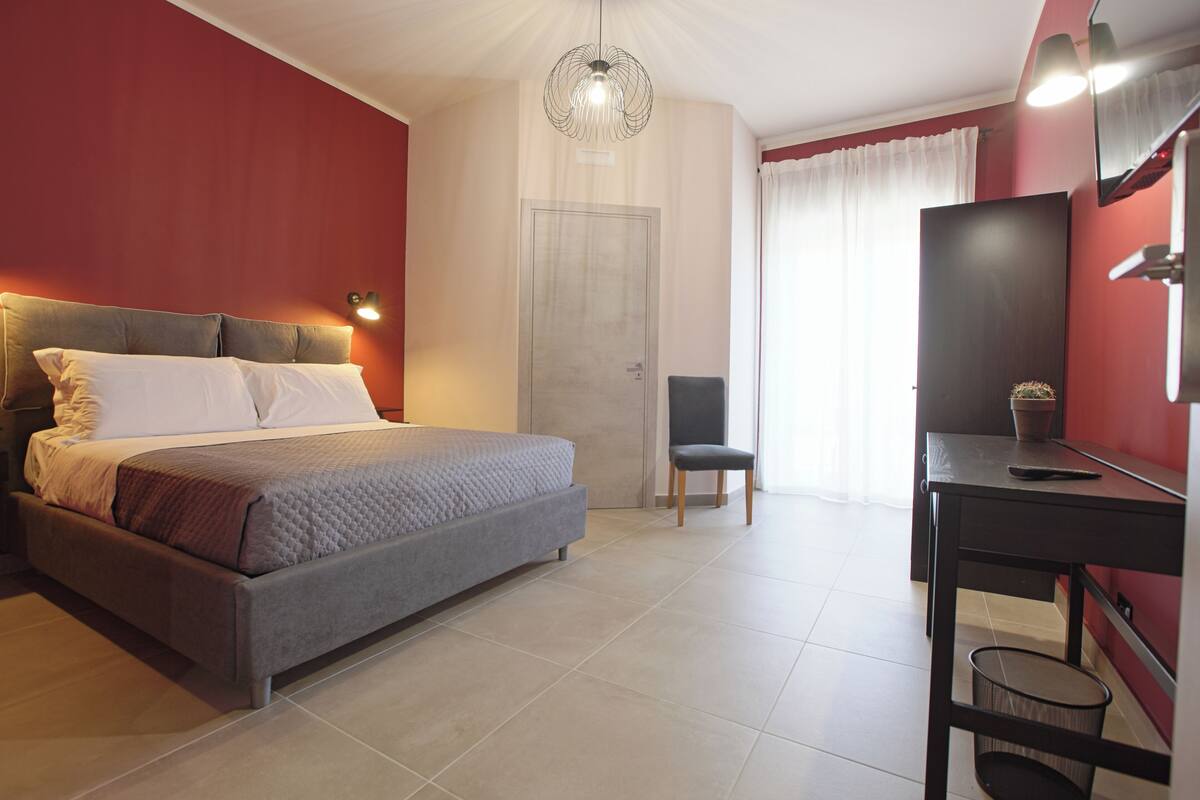 A bedroom with a bed, a desk, a chair, a door, and a hanging light fixture.