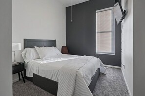 A bedroom with a bed, bedside table, lamp, and window with blinds. - Modern St. Louis Retreat – Near Downtown & Soulard (St. Louis)