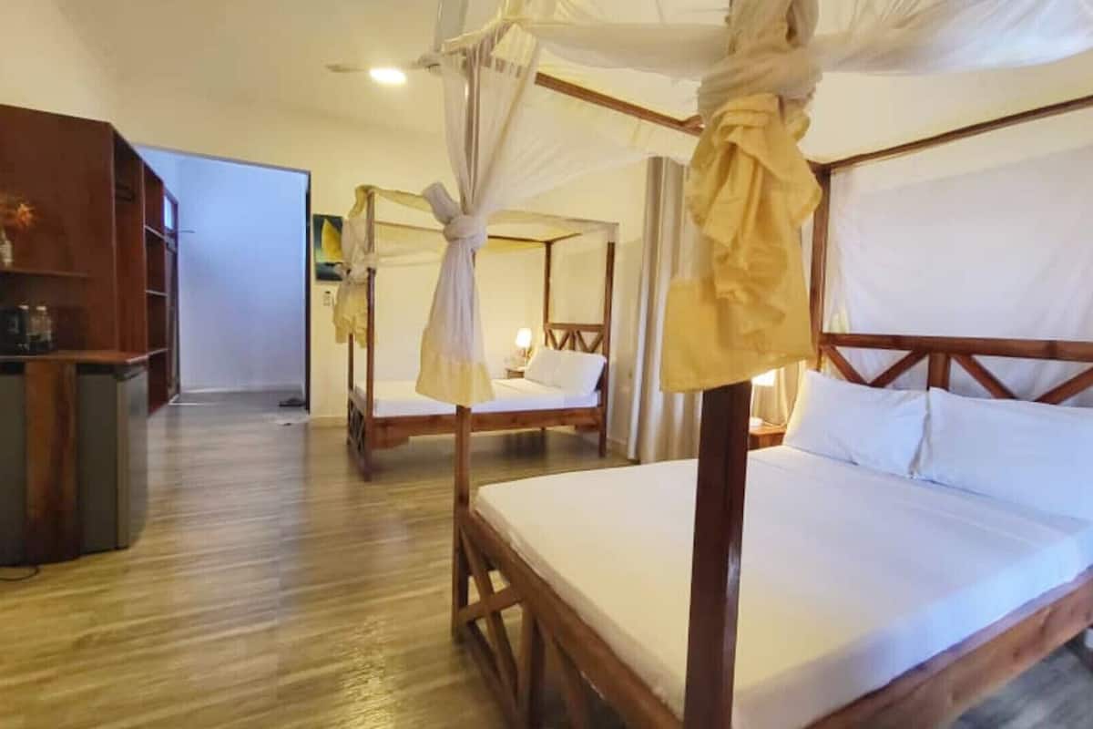 Habitación de hotel con una cama con dosel de madera, una cama individual con dosel, un armario de madera y un escritorio con lámpara.