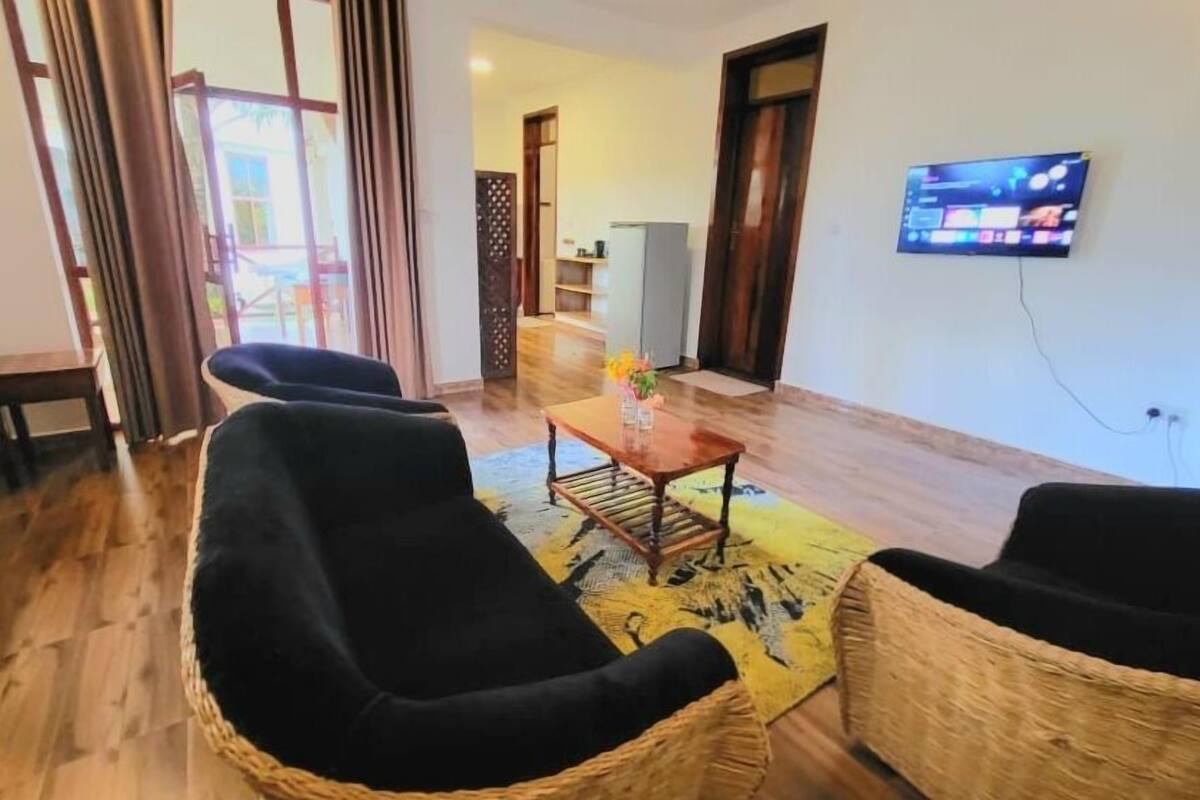 Una sala de estar con piso de madera, dos sillones negros, una mesita con un florero y un televisor montado en la pared.