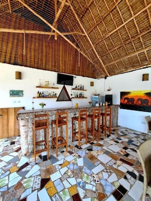 A bar area with a mosaic floor, wooden bar stools, and a television mounted on the wall. - Mimi na Wewe Bungalows Hotel (Nungwi)