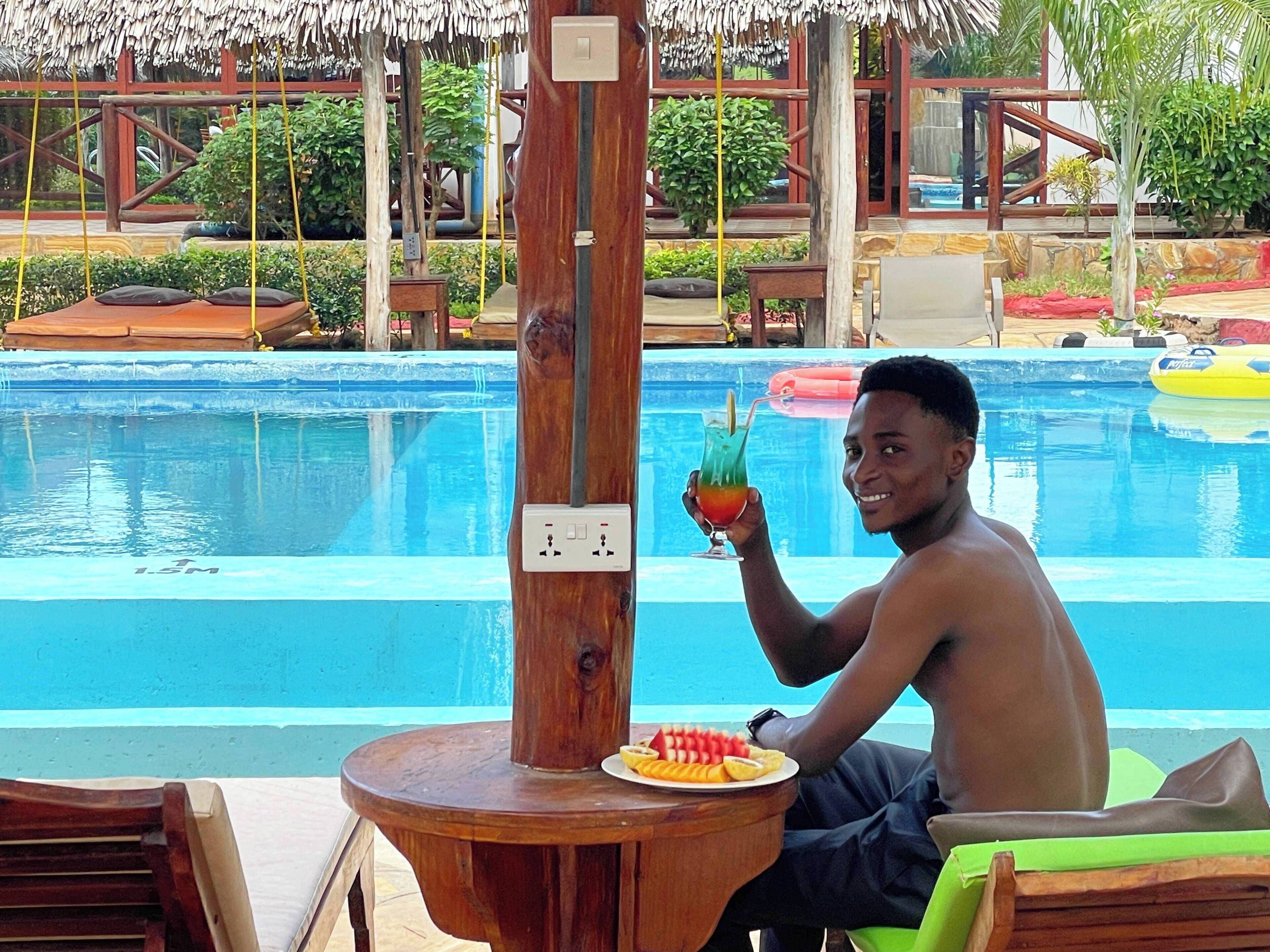 Una persona sentada junto a una piscina, con una bebida y un plato de fruta, rodeada de mobiliario de madera y una estructura de techo de paja.