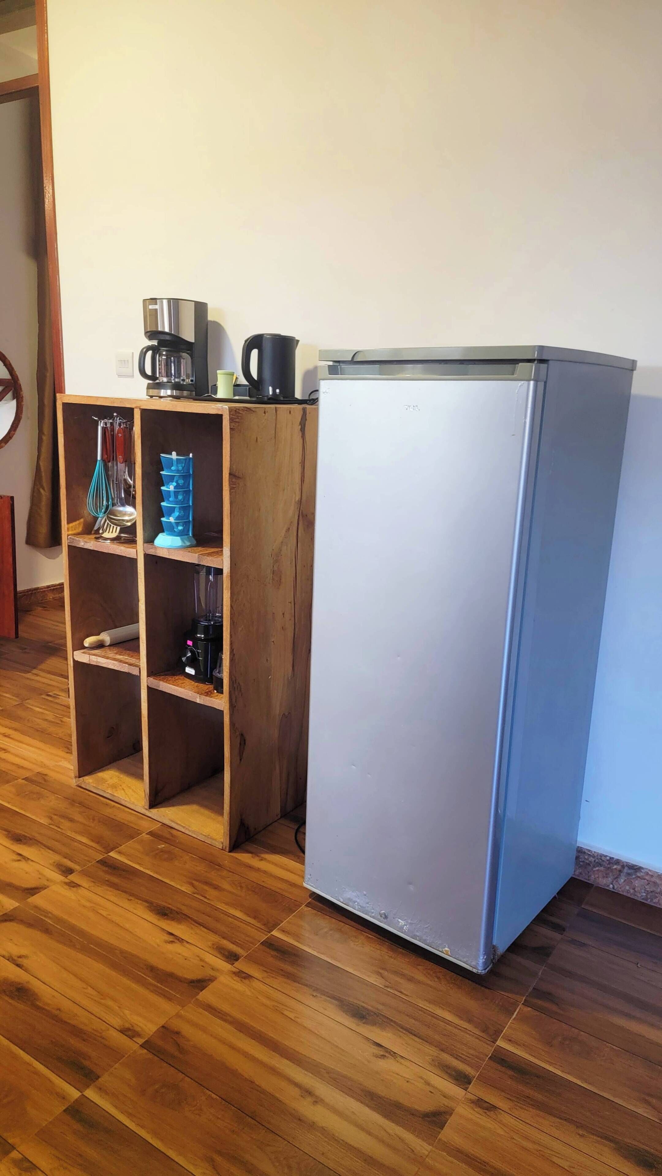 Une cuisine compacte équipée d’un réfrigérateur, d’étagères en bois, d’une machine à café et d’un mixeur.