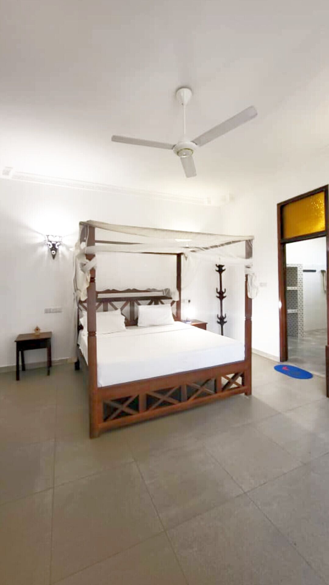 A four-poster bed with a ceiling fan, a small bedside table, and a wall-mounted light fixture.