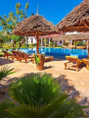 Zona de piscina con sombrillas de paja, sillas de descanso y una piscina azul cristalina.