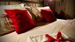 A bed with red and beige pillows, a white towel with a red ribbon, and a stone wall in the background. - Vatican Living Rome (Rome)