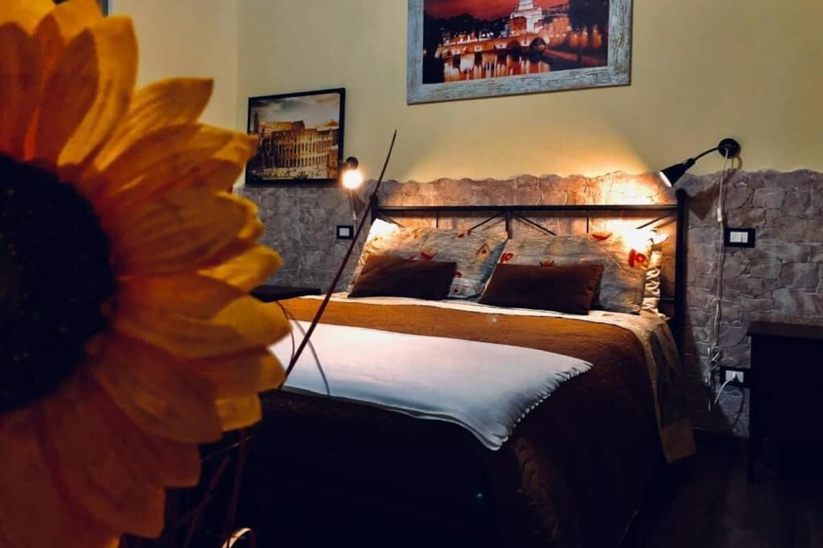 A bedroom with a stone wall, a bed with a white sheet, a framed picture above the bed, and a large sunflower in the foreground.