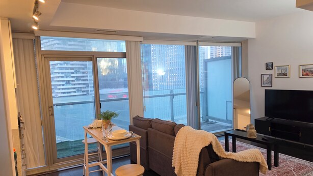 Una sala de estar con un sofá, un televisor, una mesa con una planta y ventanas amplias que dan a edificios.
