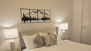 Una cama bien hecha con un edredón acolchado, dos lámparas de noche, una obra de arte en la pared y una puerta al fondo.