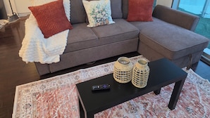 Una sala de estar moderna con un sofá seccional gris, dos almohadones rojos, una manta blanca, una mesa de centro negra con dos cestos tejidos y un control remoto.