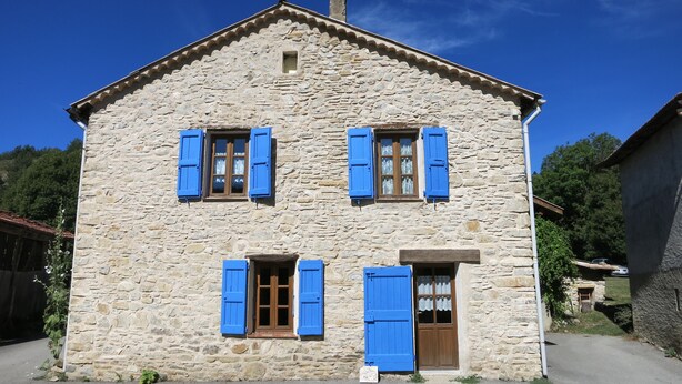 Un bâtiment en pierre doté de deux fenêtres et d’une porte, chacune étant pourvue de volets bleus.