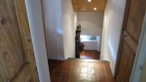 Un couloir étroit aux murs en bois et au sol carrelé mène à une chambre à coucher équipée d’un lit et d’une table de chevet.