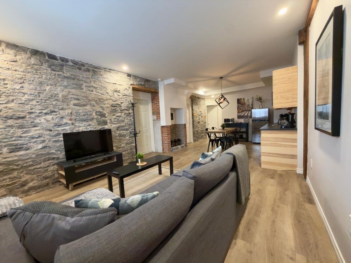 A modern living room with a stone wall, a sofa, a TV, and a kitchen area in the background.