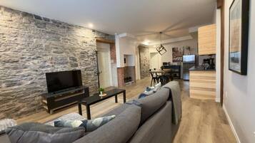 A modern living room with a stone wall, a sofa, a TV, and a kitchen area in the background.