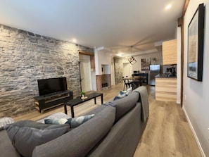 A modern living room with a stone wall, a sofa, a TV, and a kitchen area in the background. - A lovely apartment with Victorian charm (Québec)