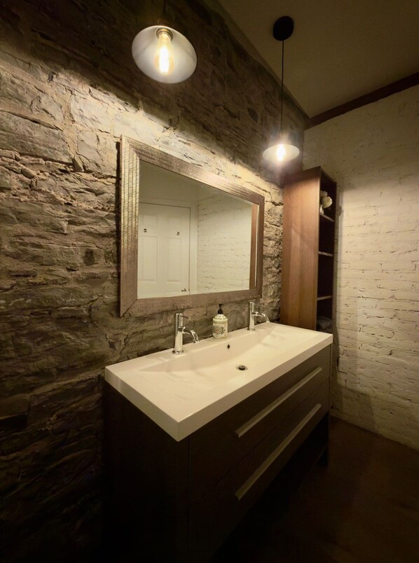 A bathroom with a stone wall, a large mirror, a double sink, and a wooden cabinet. - Charm & Prime Location | Downtown Loft (Québec)
