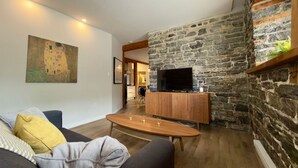 Uma sala de estar com parede de pedra, televisão de tela plana, mesa de centro de madeira e sofá com almofadas.